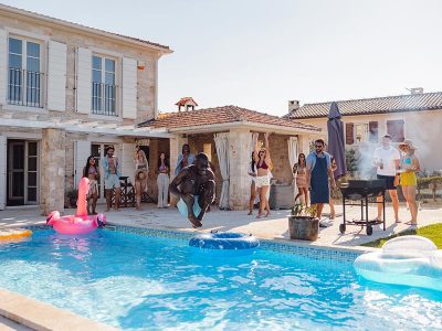 A playful adult black male jumping into the empty pool at a fun summer pool party. He is the first one to jump into the pool. His friends are watching him as he jumps. Diverse friends hanging out by the pool, grilling meat, and drinking beer. Fun weekend activities for adult friends.